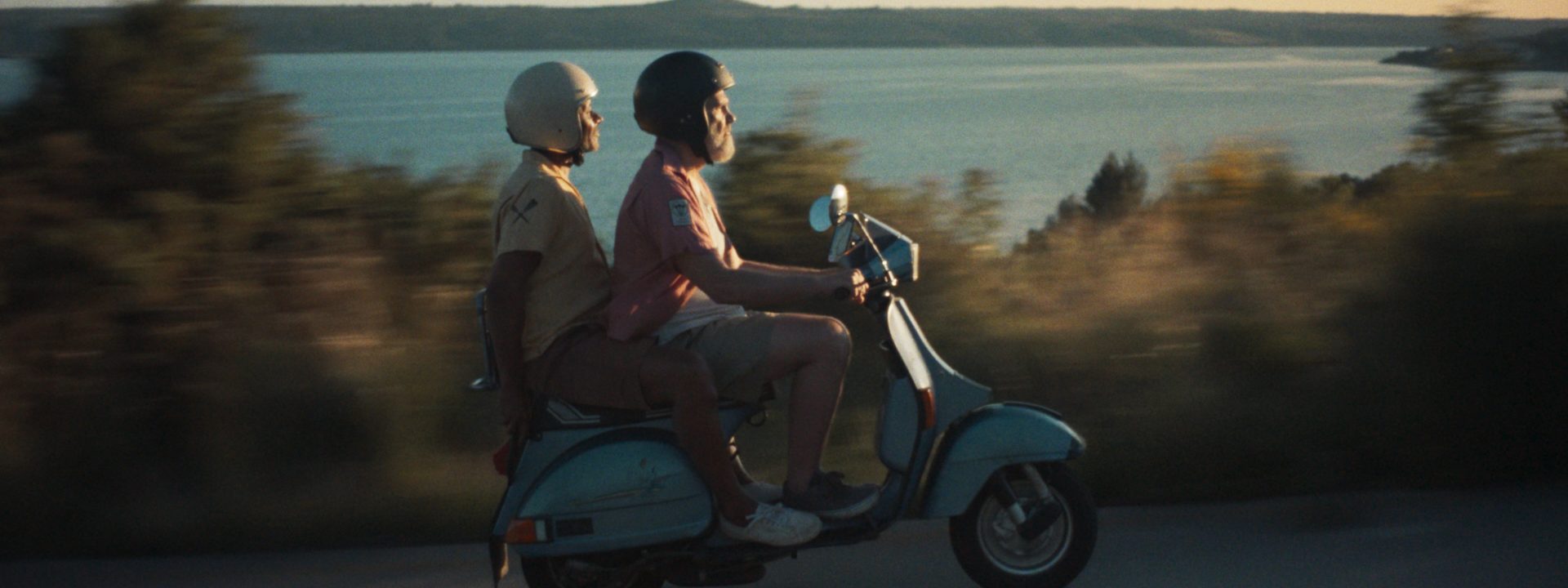 Two older men ride a vintage blue moped along a coastal road at sunset, capturing the freedom of spontaneous travel.