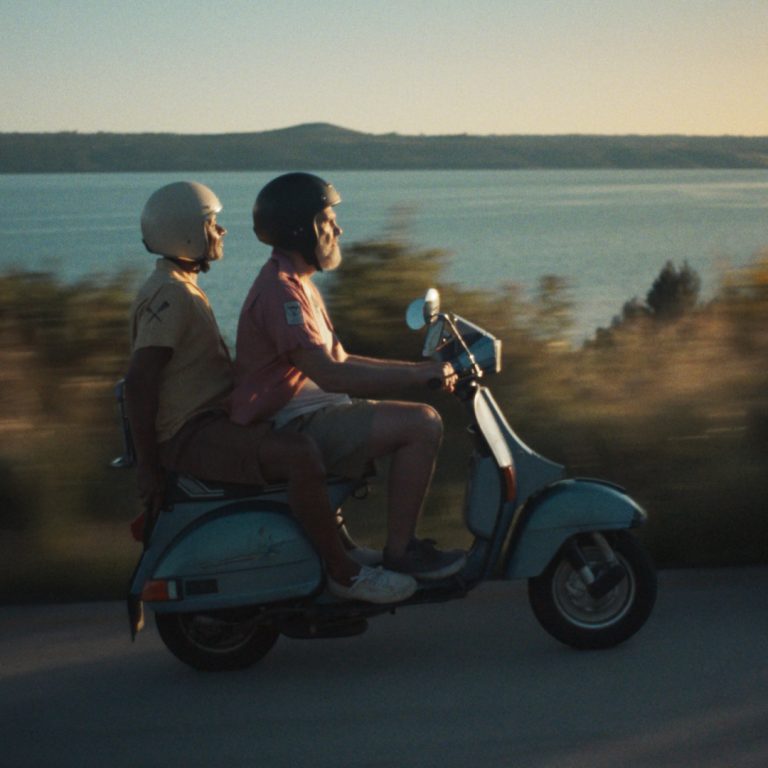 Two older men ride a vintage blue moped along a coastal road at sunset, capturing the freedom of spontaneous travel.